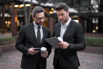 Two businessmen discussing using tablet on coffee break outdoor. Two business people talk project strategy. Two american businessmen in suits walk outdoors in the city and discuss business.