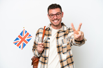 Wall Mural - Young hispanic man holding an United Kingdom flag isolated on white background smiling and showing victory sign