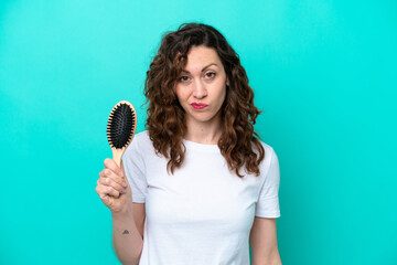 Wall Mural - Young caucasian woman holding hairbrush isolated on blue background with sad expression