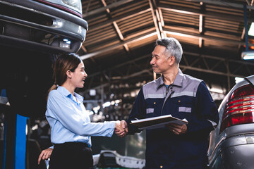 Young female car service manager giving quotation on a clipboard to senior male client and customer for his car maintenance and repair while standing in garage with vehicles for repair