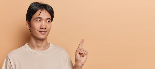 Wall Mural - Horizontal shot of handsome brunet Asian man with short hair points index finger on blank space shows promo banner dressed in casual t shirt isolated on brown background. Place your advertisement here