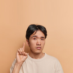 Wall Mural - Vertical shot of serious Japanese man with dark hair points index finger above advertises something asks to look at ceiling dressed in casual t shirt isolated over brown wall. Advertisement concept.