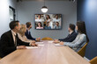 © fizkes - Engaged business team talking on online video conference call to multiethnic partners, sitting at large negotiation table in small office room, looking at display with diverse head shots