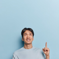 Wall Mural - Vertical shot of handsome cheerful Asian man shows advertisement demonstrates promo banner overhead smiles toothily dressed in casual t shirt isolated over blue background copy space for your text