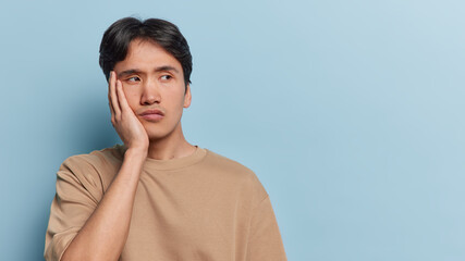 Wall Mural - Photo of sad bored male student with eastern appearance keeps hand on cheek looks unhappily aside feels tired of studying all day isolated over blue background copy space for your advertisement