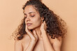 © Wayhome Studio - Half naked young woman with natural curly hair touches face gently keeps eyes closed has healthy smooth skin after beauty treatments isolated over brown background. Beauty and wellness concept