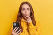 © WHstudio Leushin N - Photo of shocked woman with dark straight hair keeps mouth opened holds smartphone reacts to something unbelievable dressed in casual sweatshirt isolated over yellow background. People and reactions