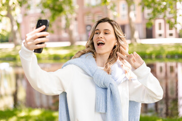 Poster - Image of smiling young curly hair blonde woman taking selfie photo holding her phone while walking in park near river. Girl make peace gesture, show two fingers, look happy and smiling.