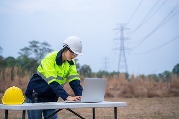 A surveying female electrical engineer wearing glasses is using a laptop computer and planning the construction of a high-voltage pylon.