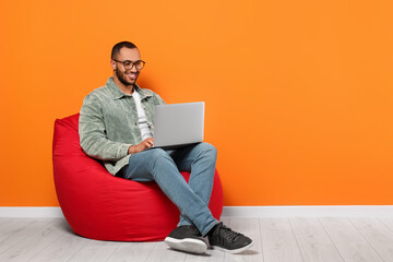 Poster - Smiling young man working with laptop on beanbag chair near orange wall, space for text