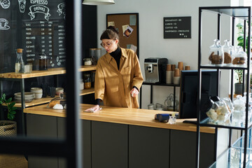 Poster - Young female clerk or owner of cozy cafe cleaning counter desk in the morning while spraying sanitizer and wiping it with duster