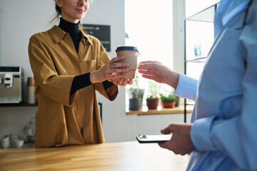 Poster - Close-up of young cafe clerk in uniform passing two cups of coffee to mature female guest over counter desk while serving her