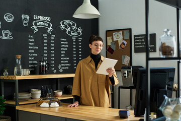 Poster - Young smiling clerk in uniform looking through online orders on tablet screen while standing by counter desk against board with menu