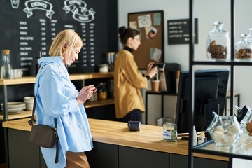 Poster - Side view of mature blond woman texting in smartphone while standing by counter desk and waiting for coffee being prepared by clerk