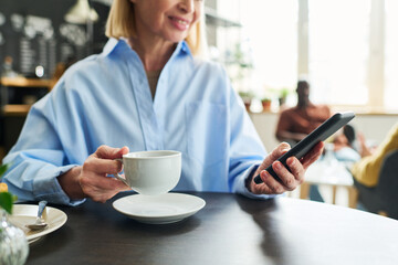 Aufkleber - Hands of mature woman holding cup of coffee and smartphone while sitting by table in cafe and texting or communicating in video chat