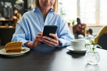 Aufkleber - Close-up of mature female consumer using smartphone by table served with cup of tea and piece of cake while having breakfast in cafe