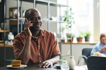Poster - Happy African American man with smartphone by ear talking to someone while sitting by served table in cozy cafe and having tea with cake