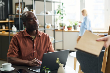 Poster - Mature businessman looking at waitress with notepad while sitting by table in front of laptop, typing and ordering something for lunch