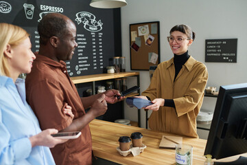 Poster - Young smiling clerk with payment terminal looking at intercultural mature couple standing by counter desk and paying for two cups of coffee