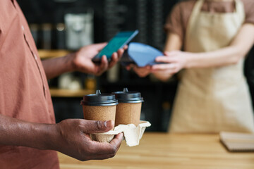 Poster - Close-up of African American mature male customer holding two cups of coffee and paying for his order by smartphone by counter desk