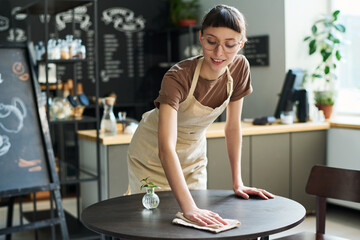 Poster - Young smiling waitress in apron bending over small round table and cleaning it with wipe while preparing it for new guests of cafe