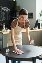 Poster - Young brunette woman in apron spraying sanitizer on surface of table and cleaning it with wipe while preparing place for new clients of cafe