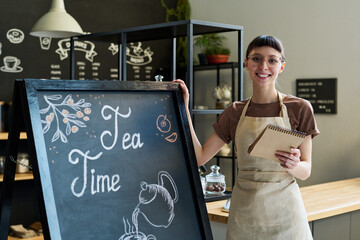 Poster - Happy young owner of cozy cafe looking at camera while standing by blackboard with advert of coffee shop drawn by white chalk