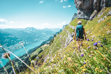 back view of mountaineer hiking on flowery trail next to steep rock wall with panoramic view on wale