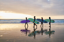 Four Surfer Friends Carrying Their Surfboards As They Walk Along The Beach At Sunset