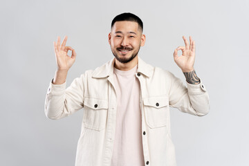 Wall Mural - Attractive smiling asian man  wearing stylish casual shirt showing ok sign, looking at camera isolated on grey  background. Positive emotions concept