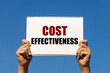 Cost effectiveness text on notebook paper held by 2 hands with isolated blue sky background. This message can be used as business concept about cost effectiveness.