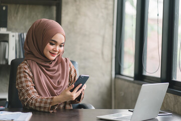 Wall Mural - Beautiful muslim business woman wearing a hijab is diligently working at her smartphone and computer, displaying professionalism and commitment to her job.