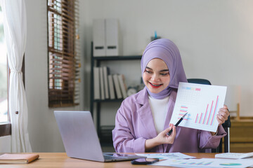 Wall Mural - A Muslim millennial businesswoman, wearing a purple hijab, is shown working with financial report paperwork in home office. The concept of financial advising, teamwork, and accounting is depicted.