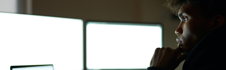 Work all night. Focused young guy sitting in dark room in front of many computer monitors while working alone late at night