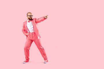 Cool young man in costume isolated on pink studio background dancing, point at copy space. Happy male in suit have fun make moves relax on party or celebration. Entertainment and fun concept.