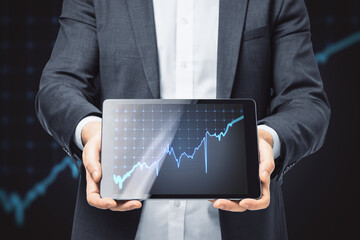 Poster - Close up of businessman hand holding tablet with growing blue financial forex chart on dark grid background. Stock, economy, market and trade concept.