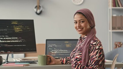 Waist up portrait of attractive young Muslim woman sitting at desk in front of computers with running program codes on monitors smiling at camera