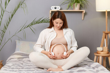 Wall Mural - Beautiful pregnant brown haired caucasian woman sitting in bed holding headphones on her tummy baby listening music future mother developing her child.