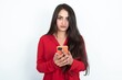 © Jihan - Portrait of serious confident Young brunette woman wearing red shirt over white studio background holding phone in two hands