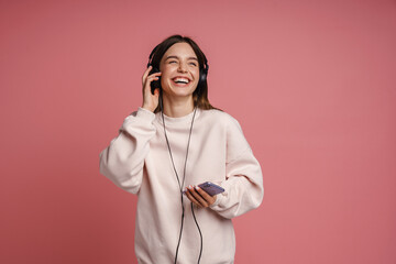 Wall Mural - Cheerful woman listening music with headphones and mobile phone isolated over pink background