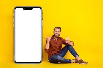 Full length portrait of cheerful person sit floor hold telephone direct thumb finger empty space board isolated on yellow color background
