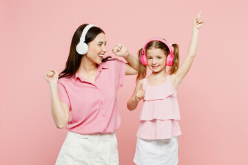Wall Mural - Happy woman wearing casual clothes with child kid girl 6-7 years old. Mother daughter listen to music in headphones, raise up hands isolated on plain pastel pink background. Family parent day concept.