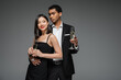smiling asian woman and elegant african american man holding champagne glasses isolated on grey.