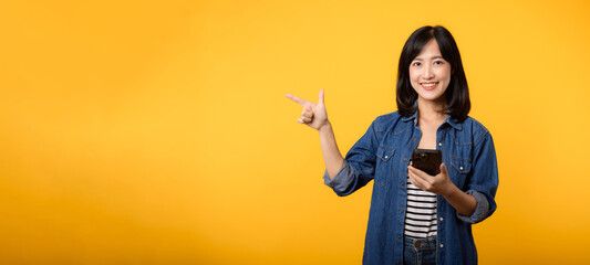 Portrait beautiful young asian woman happy smile dressed in denim jacket showing smartphone with pointing finger hand gesture to free space isolated on yellow studio background. app smartphone concept