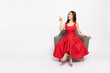 © comzeal - Happy young Asian woman in red dress sitting on armchair and pointing to empty copy space isolated on white background