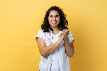 Portrait of delighted satisfied friendly woman with dark wavy hair standing looking at camera with toothy smile, clapping hands. Indoor studio shot isolated on yellow background.