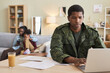 © Seventyfour - Serious military man concentrating on his online work on laptop with his family in the background