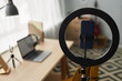 © Seventyfour - Back of smartphone in holder inside round frame standing in front of camera against workplace of student with laptop in domestic room