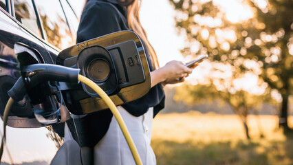 Woman charging parked electric car and browsing on smartphone