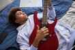 © Seventyfour - Relaxed African American teenage boy in hoodie lying on comfortable bed and playing electric guitar while enjoying leisure or weekend at home